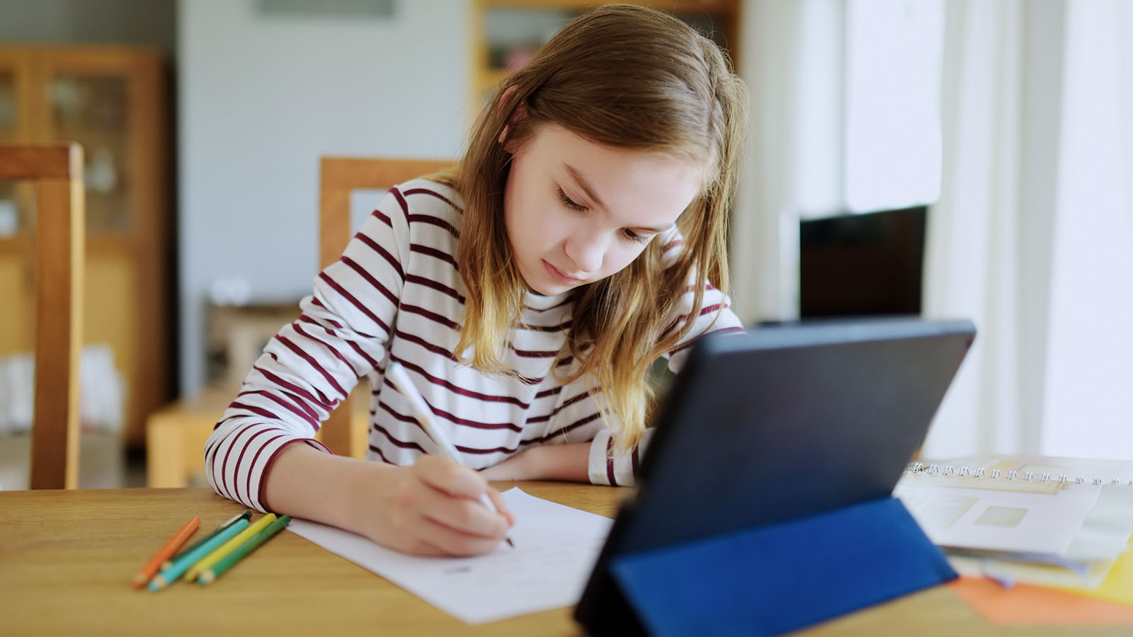 A girl of about eleven writing an answer by hand in a ruled exercise book, a tablet propped upright on a stand beside her on the same table.